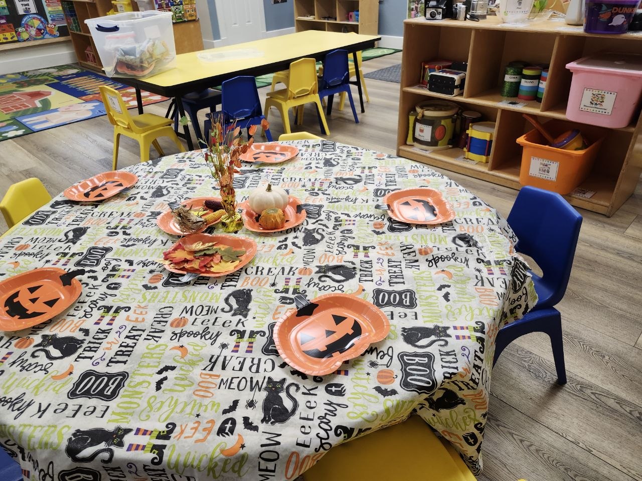 A daycare classroom decorated for Halloween, featuring a round table covered with a festive tablecloth printed with Halloween words and images like black cats and pumpkins. The table is set with orange jack-o’-lantern plates and a centerpiece made of small pumpkins, colorful leaves, and fall decorations. Bright yellow and blue child-sized chairs surround the table, with shelves of toys and art supplies visible in the background.