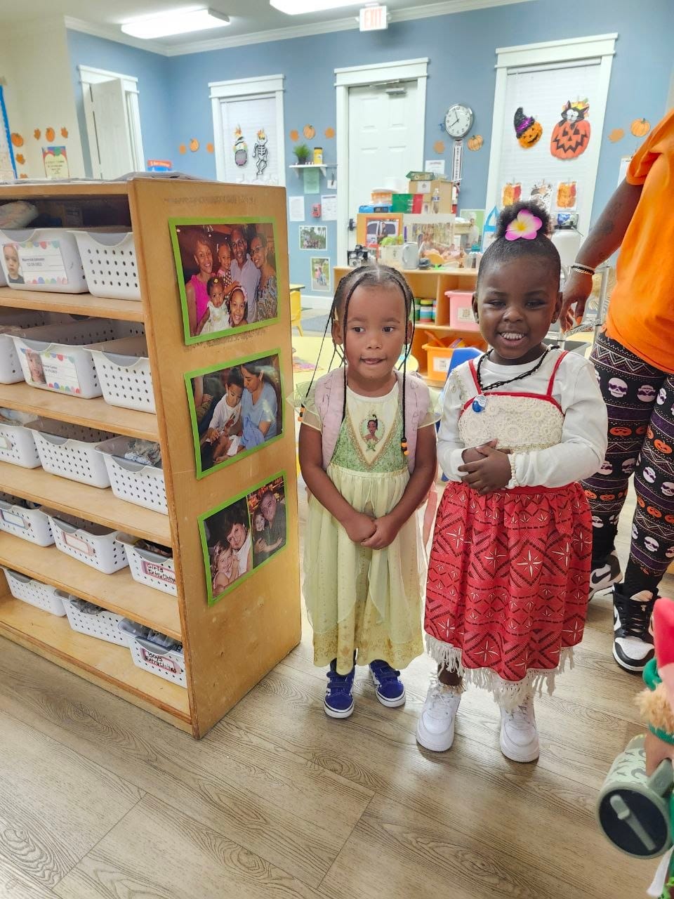 Two smiling girls standing together in a classroom, one in a pale green dress and the other in a red patterned skirt with a white top.