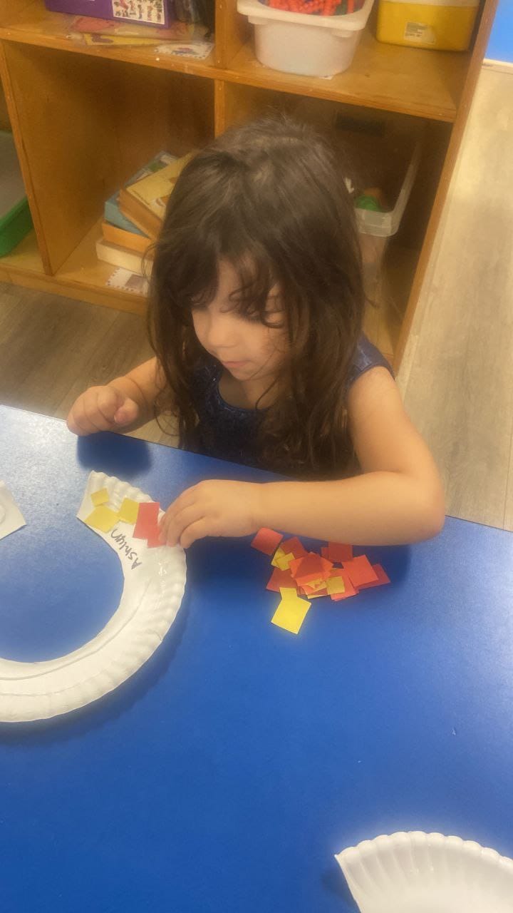A girl gluing red and yellow paper squares onto a paper plate.