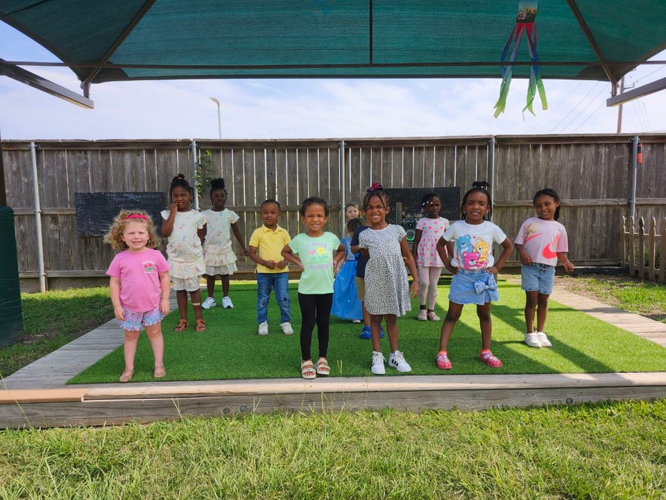 A group of children standing together on the playground turf under a shade canopy.