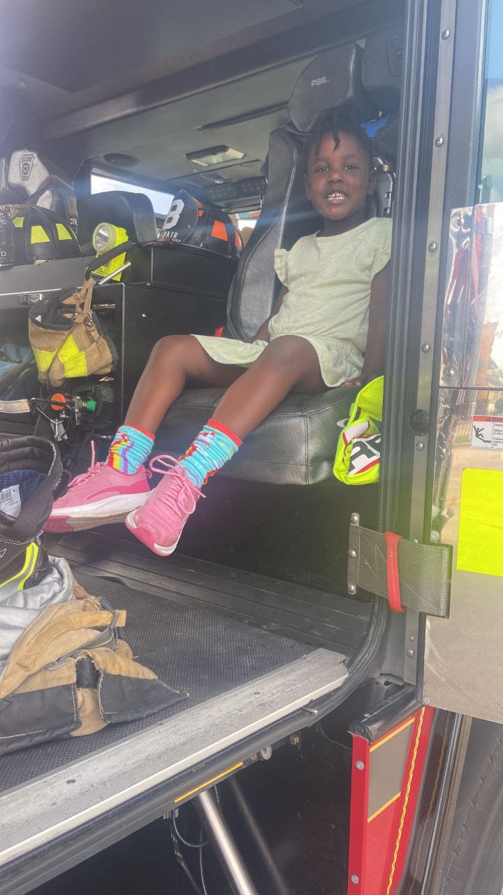 A smiling girl sitting in a firefighter's seat inside the fire truck.