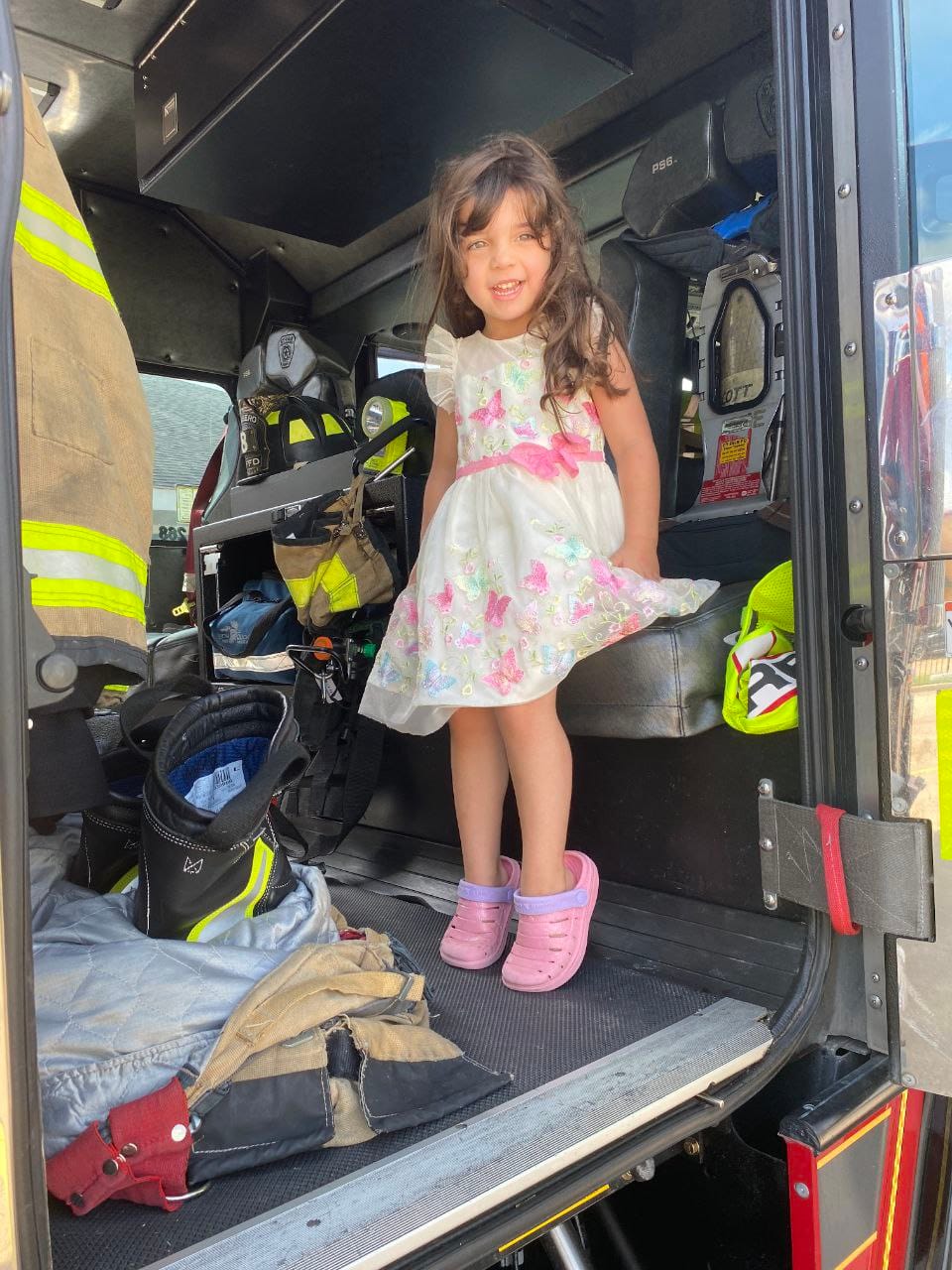 A smiling girl in a flowered dress standing inside the fire truck cab.