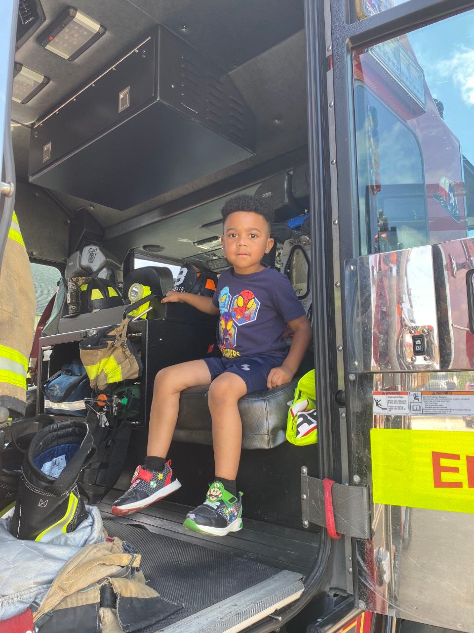 A boy in a superhero T-shirt sitting in a firefighter's seat inside the fire truck.