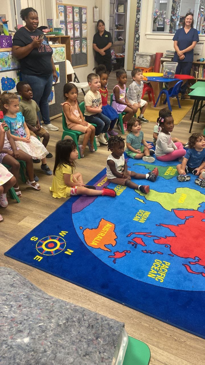 Children sitting on the classroom rug and in chairs, listening to the Science Lady as teachers stand nearby.