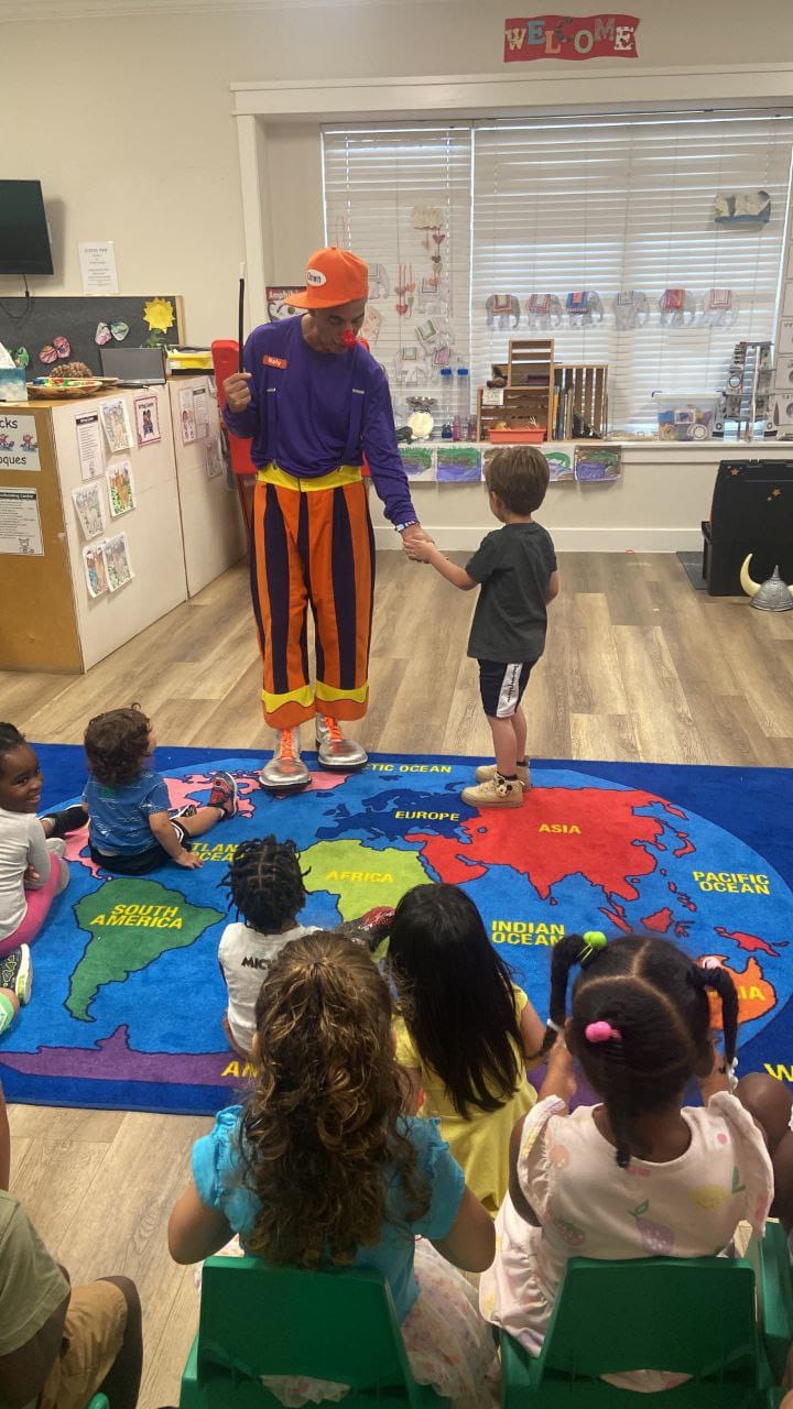 Rolly the Clown holding a boy's hand while children watch from the classroom rug.